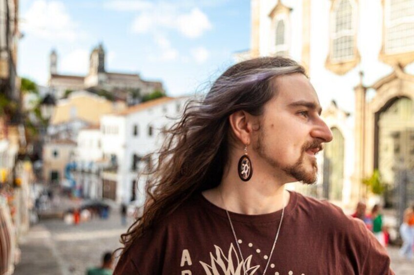 A person with long hair wearing a brown shirt and earrings, standing on a street with buildings in the background. 1