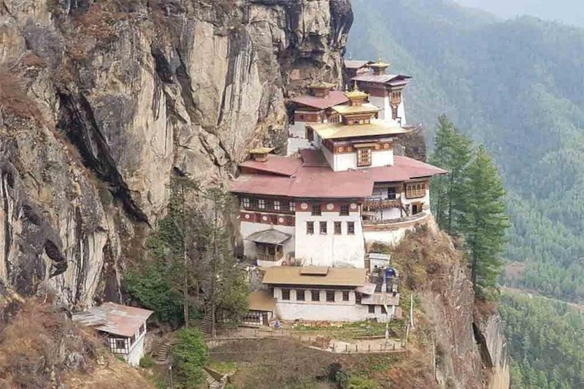 A monastery perched on a cliffside with multiple buildings, surrounded by steep rocky terrain and dense forest. 1