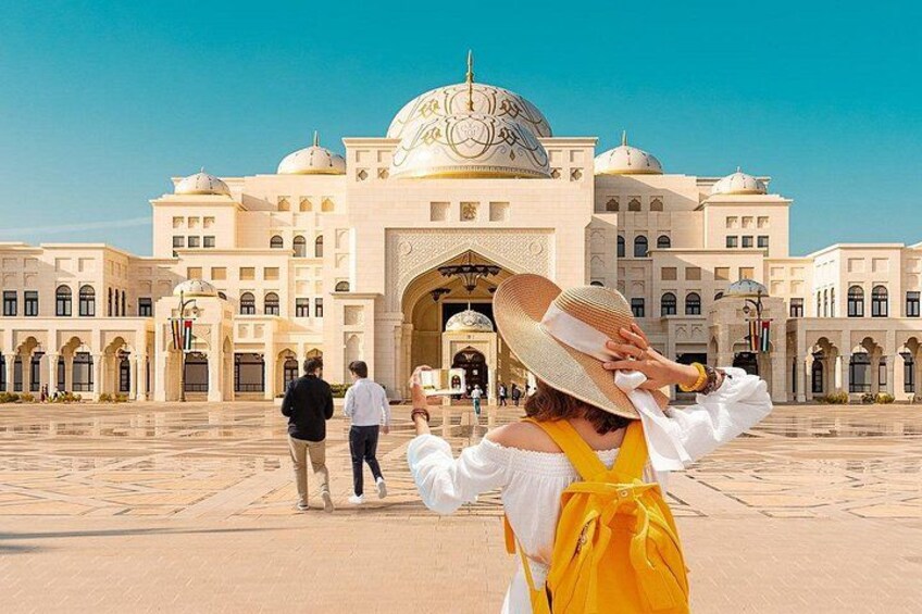 A large, ornate building with domes and arches, a paved plaza, and a few people walking. 1