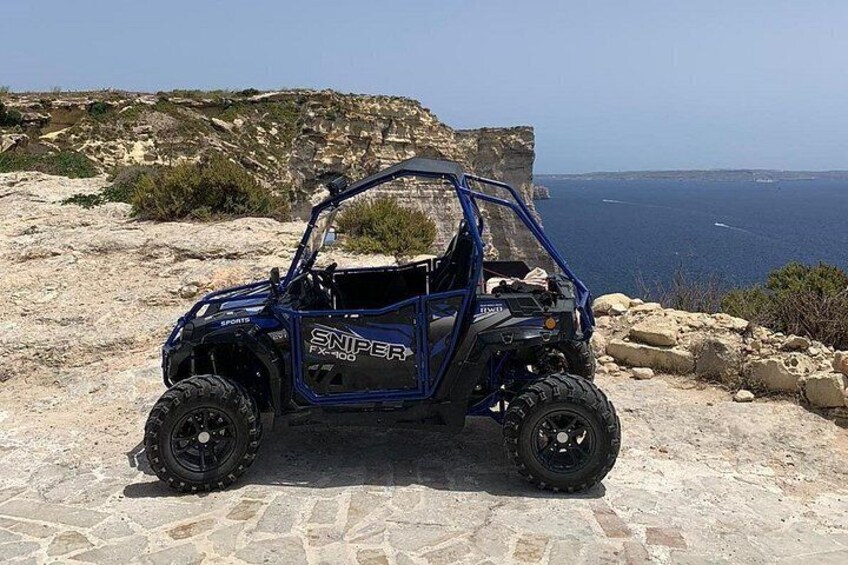 Un vehículo todoterreno azul con la leyenda 'SNIPER FX 100' en la carrocería, estacionado sobre terreno rocoso con vista al mar y acantilados al fondo. 2