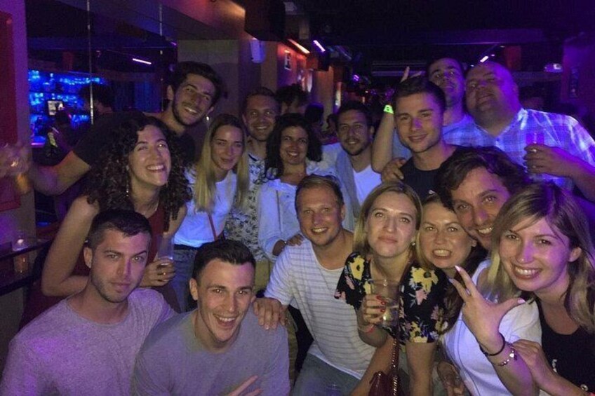 En gruppe mennesker i et dunkelt, indendørs miljø, måske en bar eller natklub, hvor mange står tæt sammen. 2