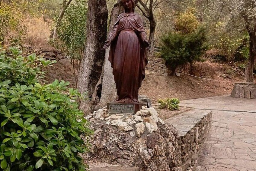 Una estatua de una mujer con vestido largo sobre un pedestal de piedra, con una placa conmemorativa, rodeada de follaje y árboles. 3