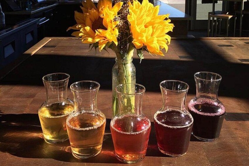 A wooden table with five glass bottles containing different colored liquids, a vase with yellow flowers, and a wooden bench in the background. 2