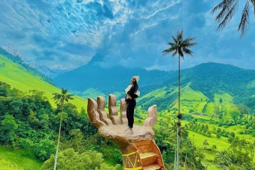 A person standing on a large hand sculpture overlooking a lush green valley with palm trees and mountains in the background. 1