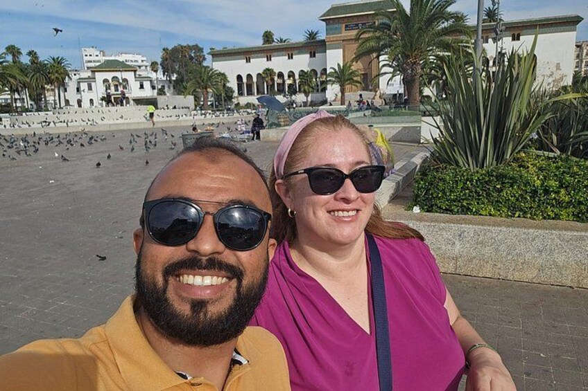 A man and a woman taking a selfie in a public square with buildings and palm trees in the background. 4