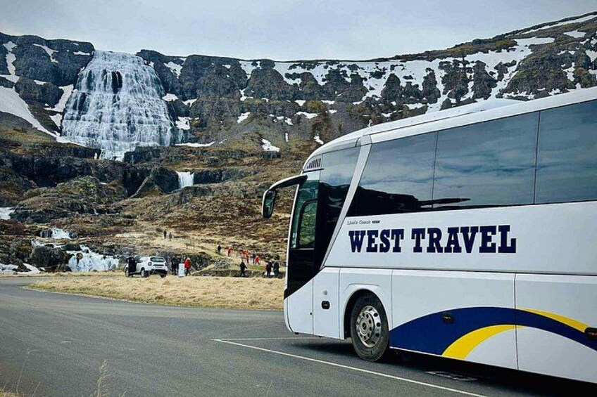 Un autobús con la leyenda 'WEST TRAVEL' en su lateral, estacionado en una carretera con paisaje montañoso y una cascada al fondo. 2
