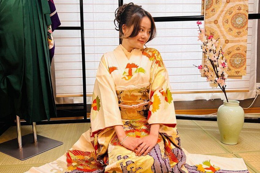 A person in traditional Japanese attire sitting on tatami flooring with a vase of flowers and a green garment in the background. 3