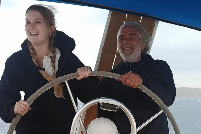 Two people steering a boat, with a clear sky and calm sea in the background. 2