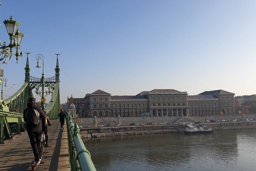 A pedestrian bridge with a green railing, a large building with classical architecture in the background, and a calm river. 3