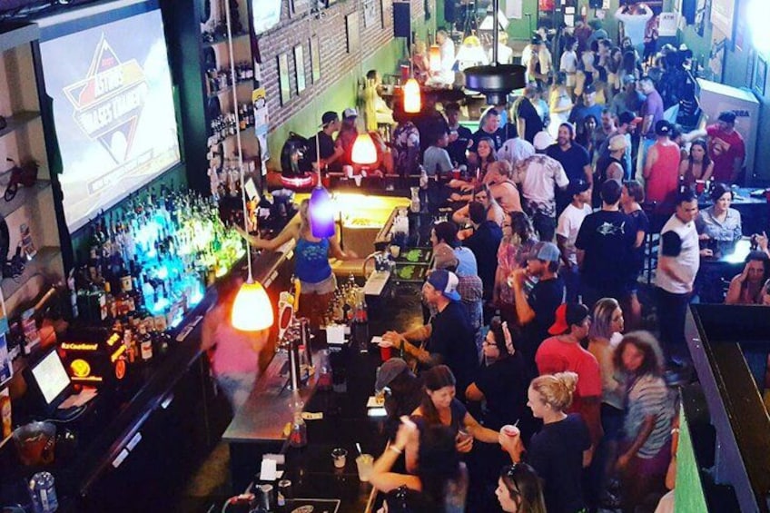 Een drukke bar met gezellige mensen, een toog vol flessen en een projectiescherm. 3