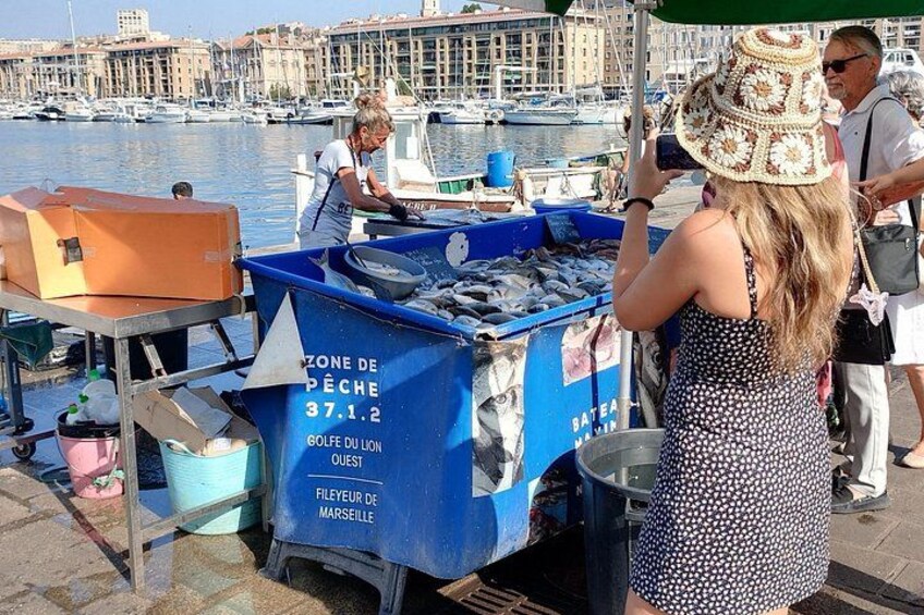 Un venditore di pesce in un mercato sul lungomare, con un carrello blu con la scritta 'ZONE DE PÊCHE 37.1.2' e 'GOLFE DU LION OUEST'. 1