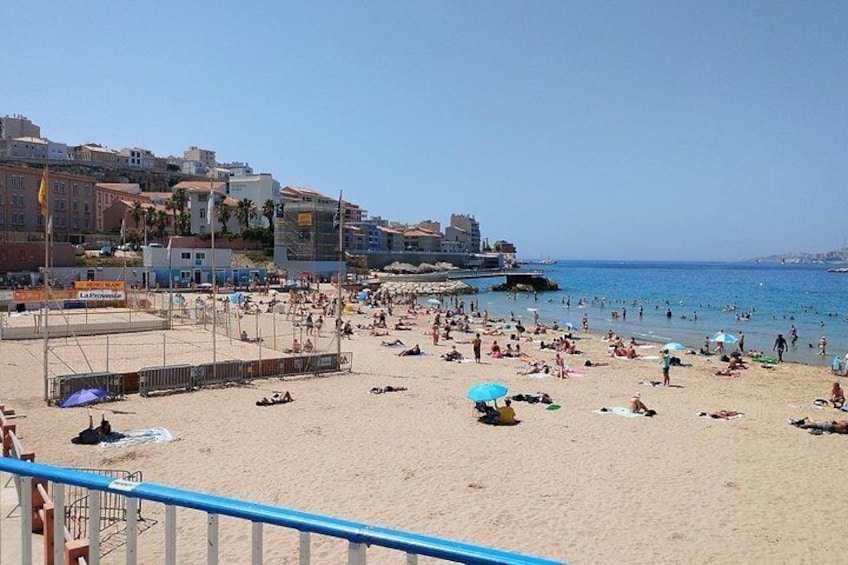 A crowded beach with people sunbathing and swimming, a clear blue sky, and buildings in the background. 5