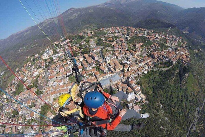 A person in a red jacket and blue helmet preparing for a paragliding flight over a town surrounded by mountains. 4