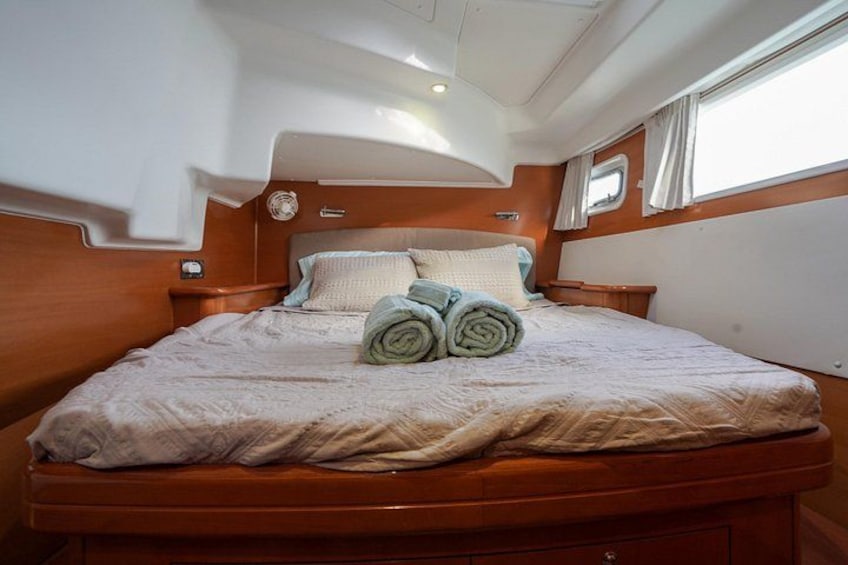 A neatly made bed with rolled towels, wooden headboard, and a small window with curtains. 4