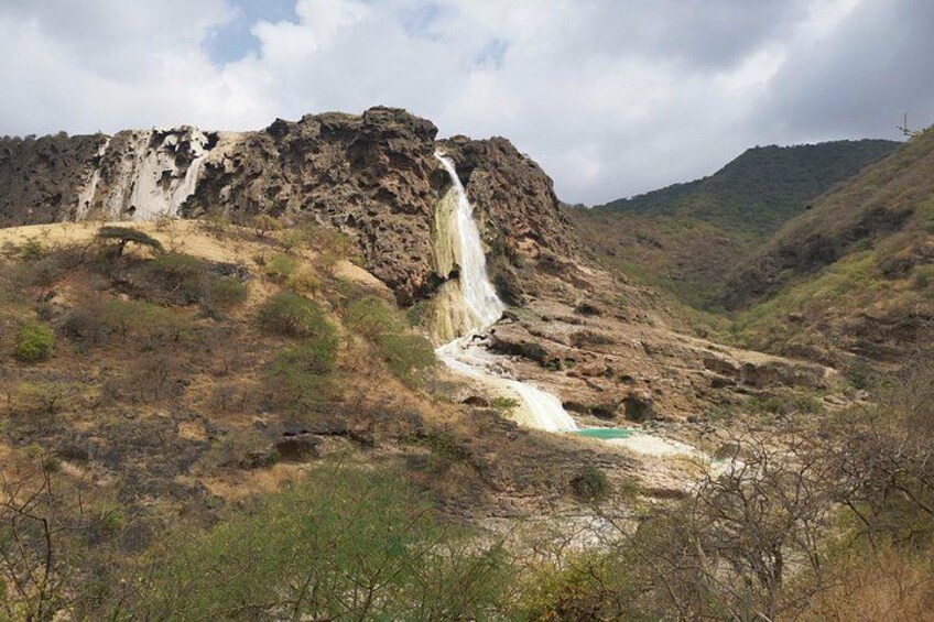Ein Wasserfall stürzt eine felsige Hangkante hinab, umgeben von trockener Vegetation und wolkenlosem Himmel. 2