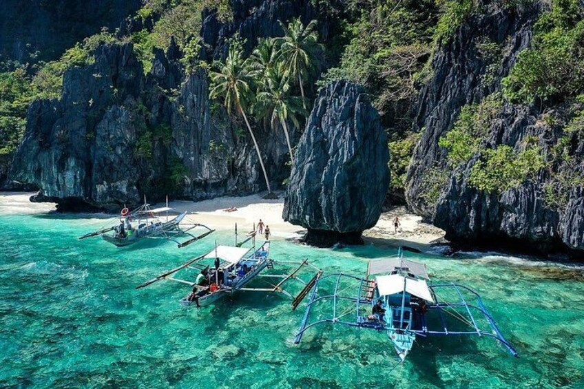 A clear turquoise sea with traditional boats anchored near a rocky coastline with palm trees. 5