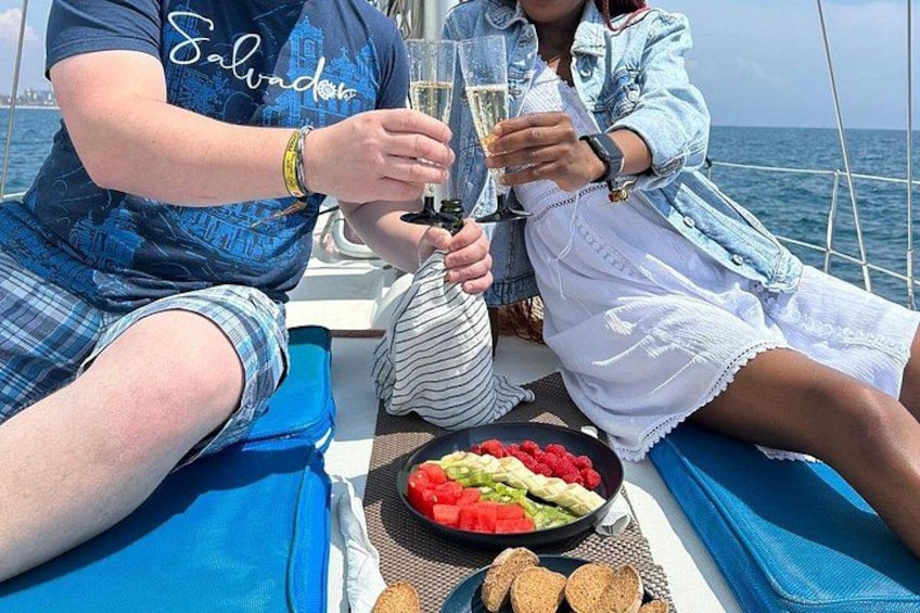 Two people on a boat, holding glasses, with a plate of food and a bottle in front of them. 3