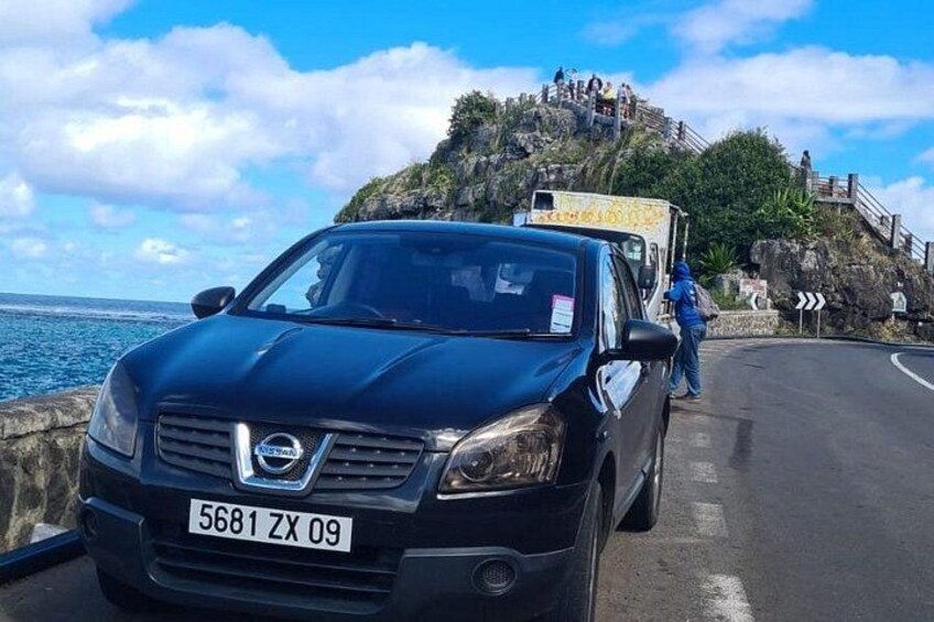 A black car parked on a coastal road with a rocky cliff and a sea view in the background. 3