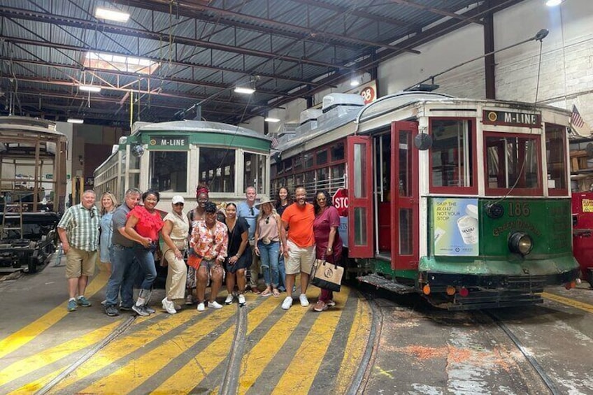 A group of people standing in front of a vintage tram labeled 'M-LINE' in a workshop or factory setting. 1