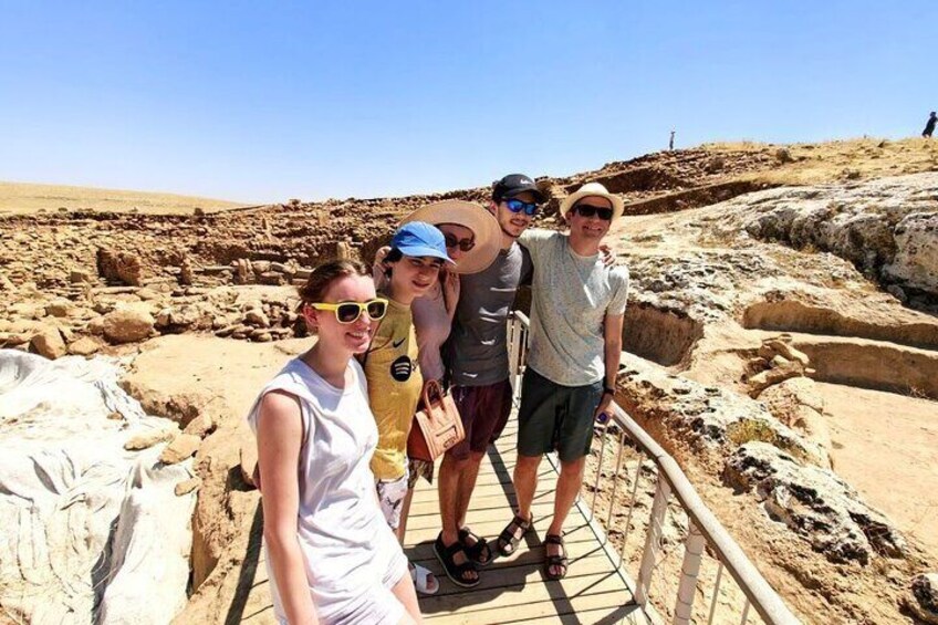 A group of people standing on a wooden platform at an archaeological site with visible excavation areas and rocky terrain. 1