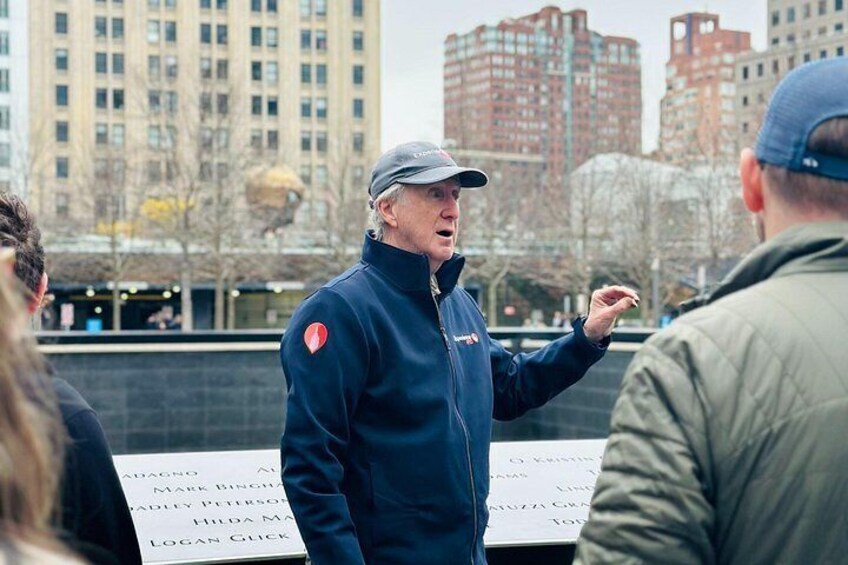 A person in a blue jacket and cap gesturing, with a memorial wall in the background. 3