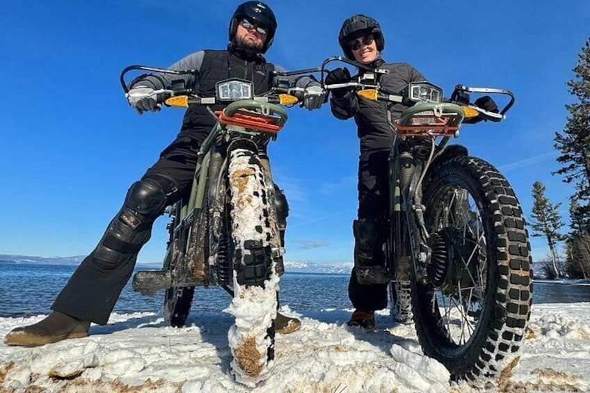 Two people on all-terrain vehicles on a snowy surface with a body of water and trees in the background. 2