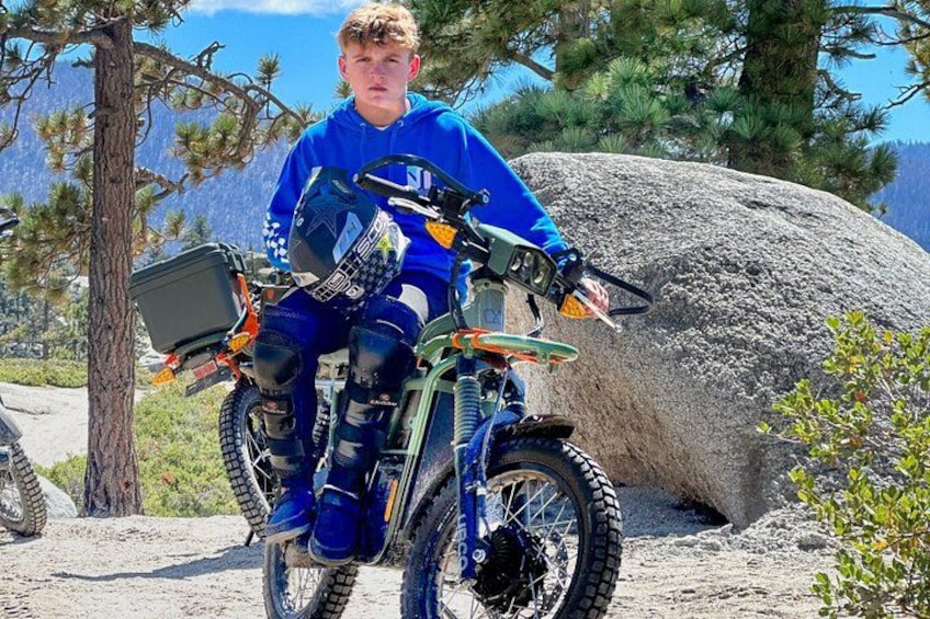 A person on a dirt bike with protective gear, a helmet, and a backpack, in a natural outdoor setting with trees and a large rock. 2