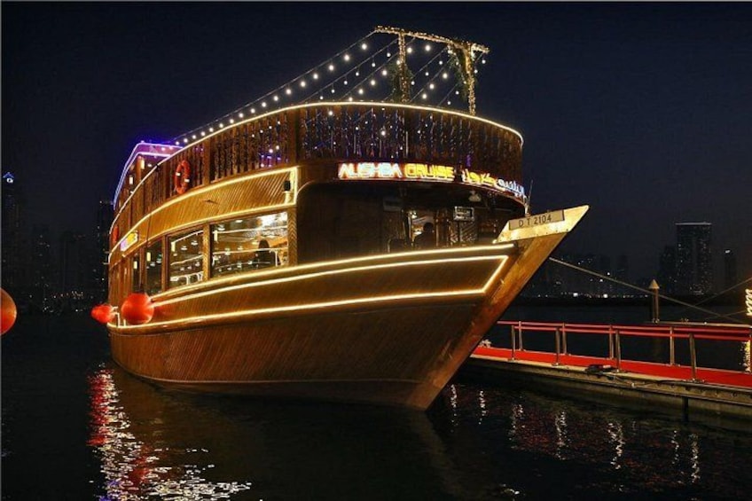 A large, illuminated boat with a deck and multiple levels, docked at a port with a city skyline in the background. 1