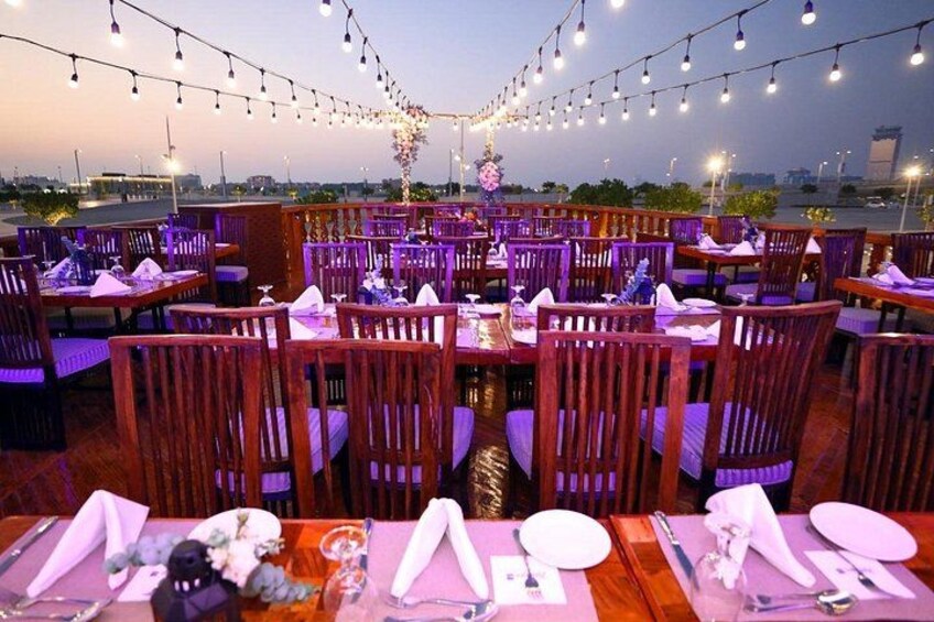 An outdoor restaurant with wooden tables and chairs, string lights, and a view of the cityscape. 2