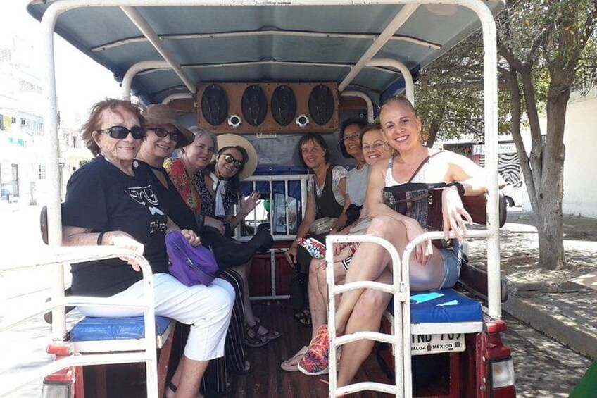 A group of people on a stationary vehicle with a covered top, some wearing hats, and a tree visible in the background. 3