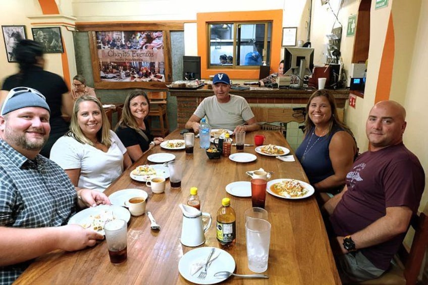 A group of people sitting around a wooden table with plates of food, drinks, and a pitcher. 4