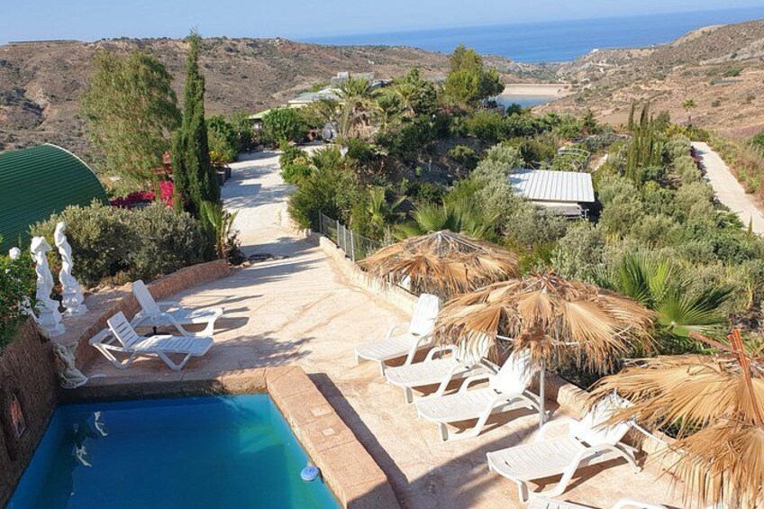 Ein Pool mit Liegestühlen, einem Reetdach-Sonnenschirm und Blick auf das Meer und die Hügel. 1