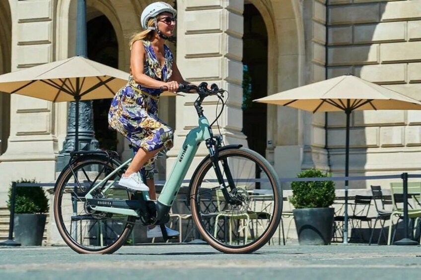 A person riding a green electric bicycle with a white helmet, in front of a building with columns and outdoor seating. 1