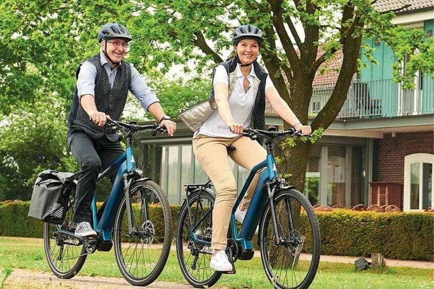 Two people riding bicycles on a sidewalk, with a house and trees in the background. 5