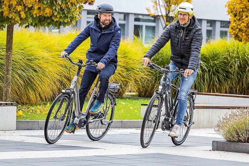 Two people riding bicycles on a paved path with greenery and buildings in the background. 2