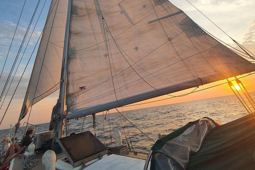 A sailboat with a person on deck, sails unfurled, and the sun setting over the water. 4