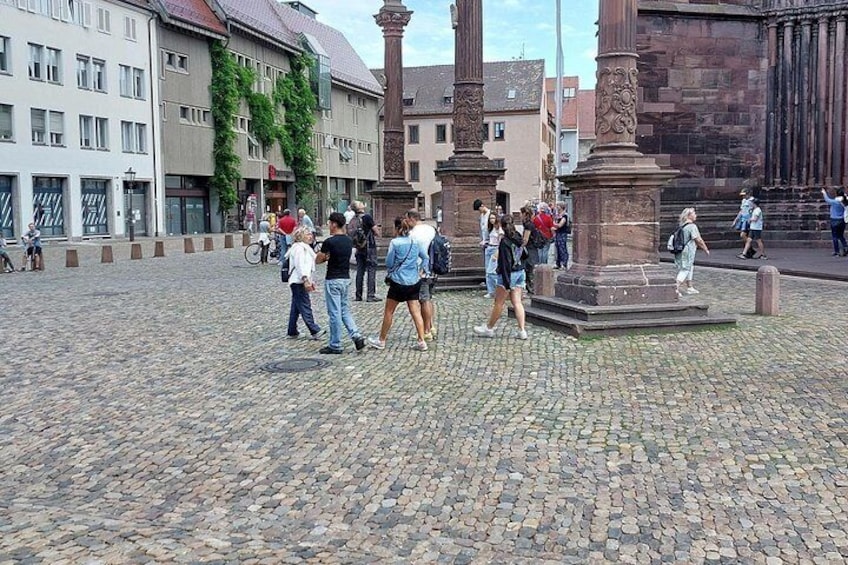 A cobblestone square with people walking, historic buildings, and a central monument. 2