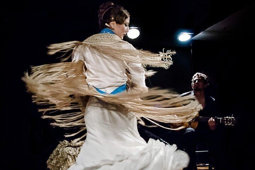 Una persona in un abito bianco fluente con dettagli blu che balla, con una chitarra sullo sfondo. 2
