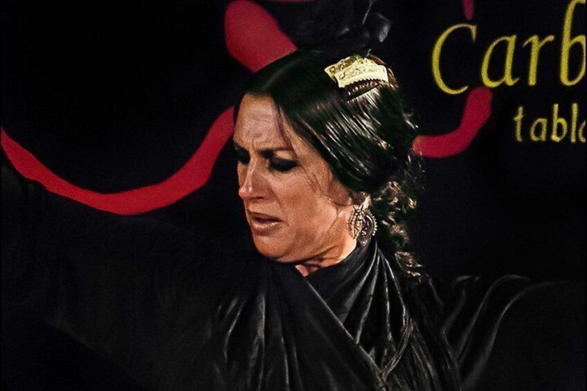 Una persona dai capelli scuri, vestita di nero, in piedi di fronte a uno sfondo con elementi decorativi rossi e neri. 3