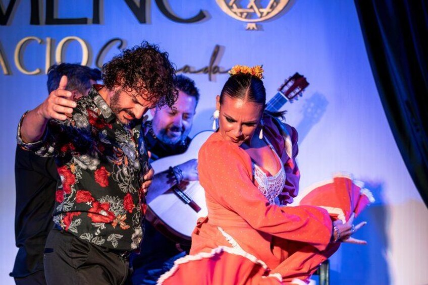 A person in a red dress with a white collar, holding a microphone, and another person in a floral shirt gesturing. 1