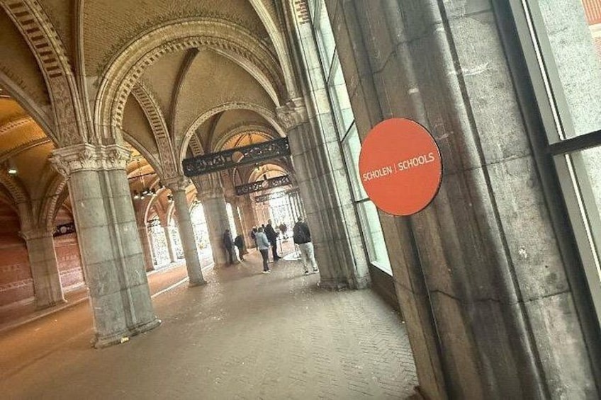 Un intérieur avec des plafonds voûtés et des colonnes, une enseigne indiquant « SCHOLEN | SCHOOLS », et des gens qui se promènent. 2