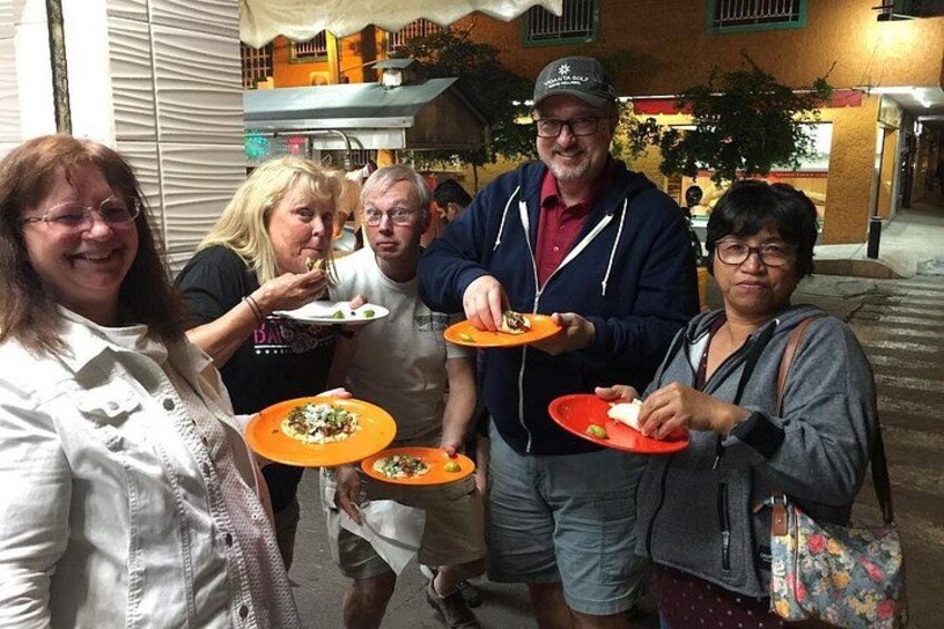 A group of people holding plates with food, standing on a street at night. 2