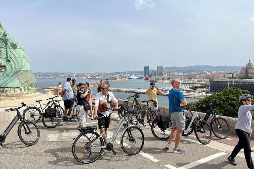 A group of people near a waterfront with bicycles, a large green sculpture, and a cityscape in the background. 5