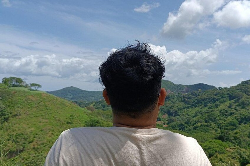 A person facing away from the camera, overlooking a hilly landscape with greenery and distant hills. 3