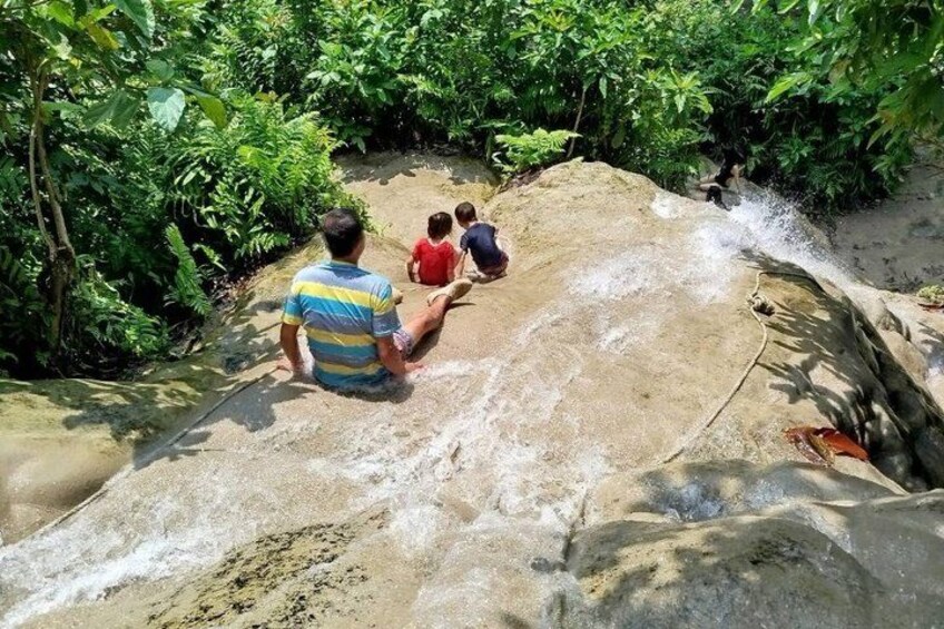 Un homme et deux enfants sont assis sur un rocher, au bord d’un ruisseau qui serpente à travers une zone verdoyante et luxuriante. 2