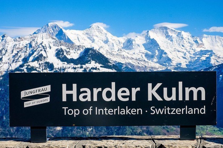 Ein Schild mit der Aufschrift 'Harder Kulm' und 'Höchstpunkt von Interlaken, Schweiz', mit einer Bergkette im Hintergrund. 1