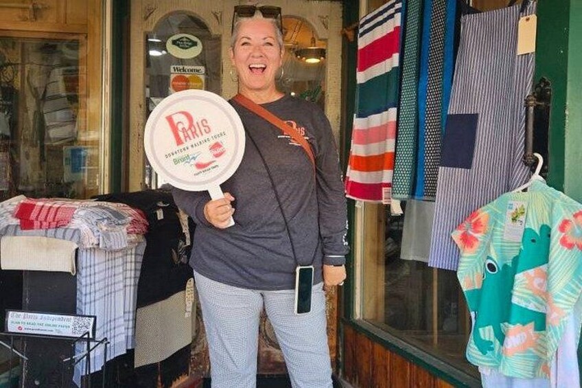 A person holding a sign with 'Paris' written on it, standing in front of a store with clothing items displayed. 2
