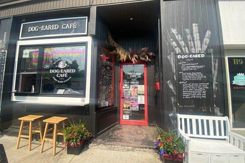 A storefront with a sign reading 'DOG-EARED CAFÉ' and a red door. There are two wooden stools outside and potted plants near the entrance. 5