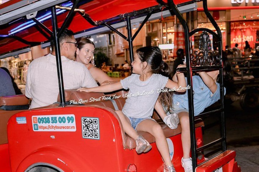 A red tuk-tuk with a QR code and a website URL on the back, a man and a woman sitting in the front, and a child in the back. 2