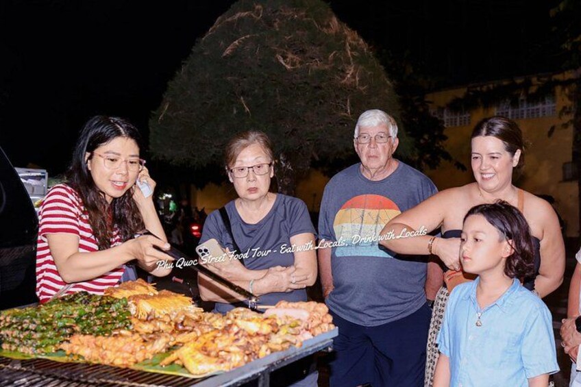 A group of people at an outdoor food event with a variety of dishes on a grill. 3